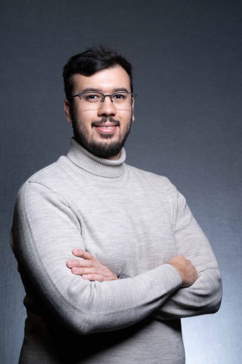 Professional headshot of a man with dark hair and glasses wearing a light gray turtleneck against a gray background, arms crossed.